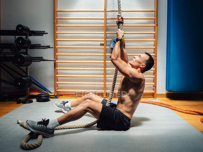 Vue latérale d'un sportif musclé torse nu montant une corde tout en faisant de l'exercice dans une salle de gym moderne — Photo de stock