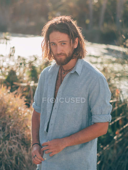 Jovem homem hipster barbudo alegre perto do rio olhando na câmera no verão ensolarado — Fotografia de Stock