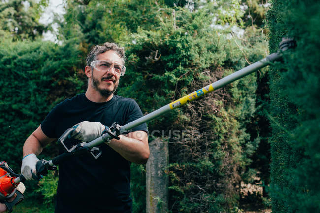 Homem caucasiano aparando uma sebe de arizonica com ferramentas mecânicas — Fotografia de Stock