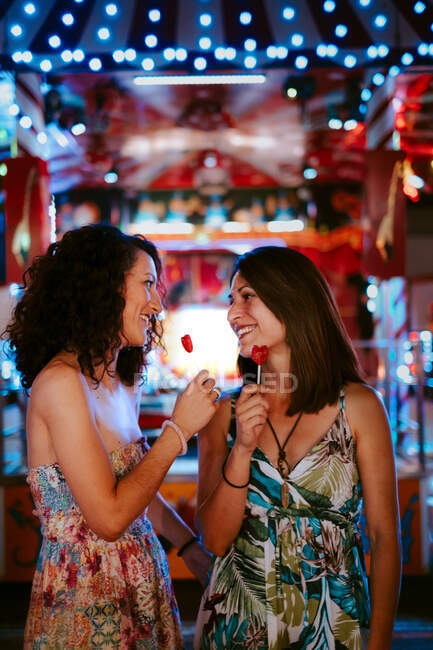 Amigos do sexo feminino com pirulitos ligação no parque de diversões na noite quente de verão olhando uns para os outros no fundo borrado — Fotografia de Stock