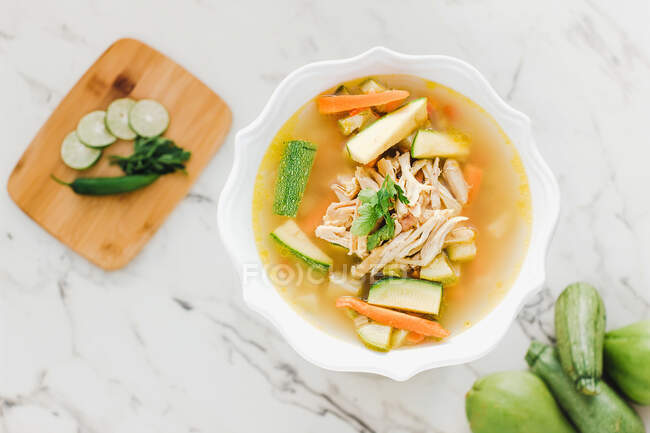 Sirve tazón de sopa de pollo con zanahoria y calabacín en la mesa con calabacín y tabla de cortar - foto de stock