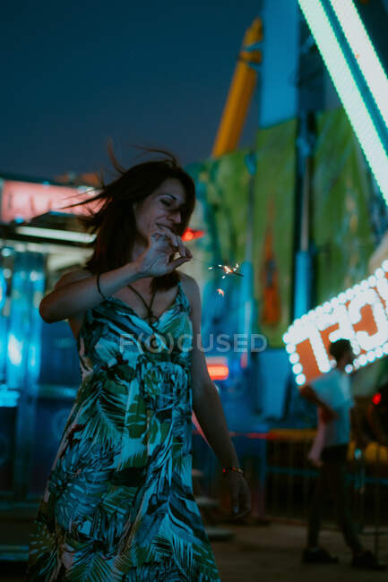 Cheerful woman wearing summer dress and spending time in amusement park holding burning sparkler on blurred background — Stock Photo