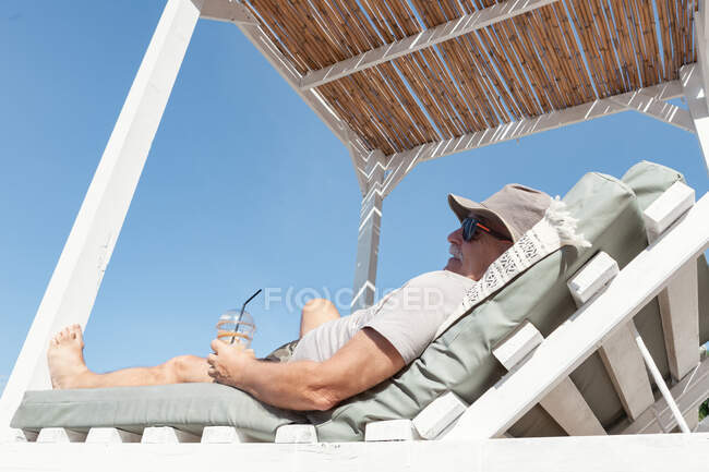Hombre bronceado con gafas de sol y sombrero disfrutando del descanso bajo la sombrilla tomando una bebida fresca en el día de verano, Halkidiki, Grecia - foto de stock