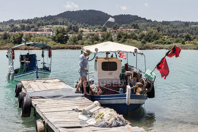 Seitenansicht einer reifen Frau, die an einem Holzsteg vorbeigeht, während sie an Bord einer bunten alten Jacht in ruhigem Wasser geht, Griechenland — Stockfoto