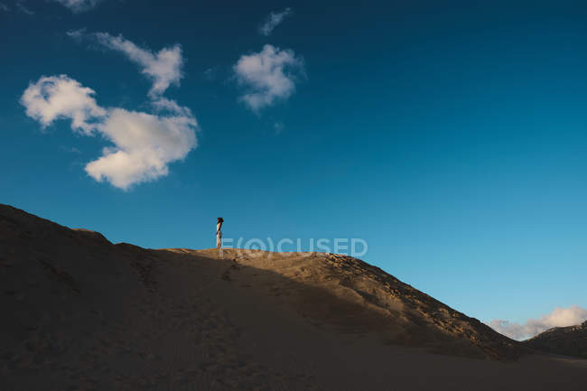 Mulher de vestido branco de pé em distante no sol iluminado colina arenosa em Tarifa, Espanha com céu azul e nuvem branca — Fotografia de Stock