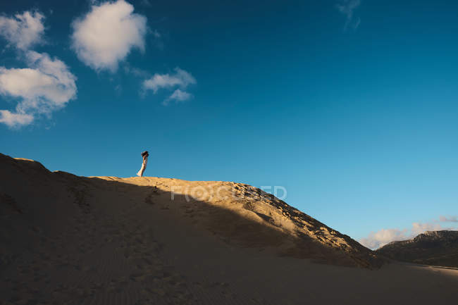 Mujer vestida de blanco de pie en la lejana colina de arena iluminada por el sol en Tarifa, Cádiz con cielo azul y nube blanca - foto de stock