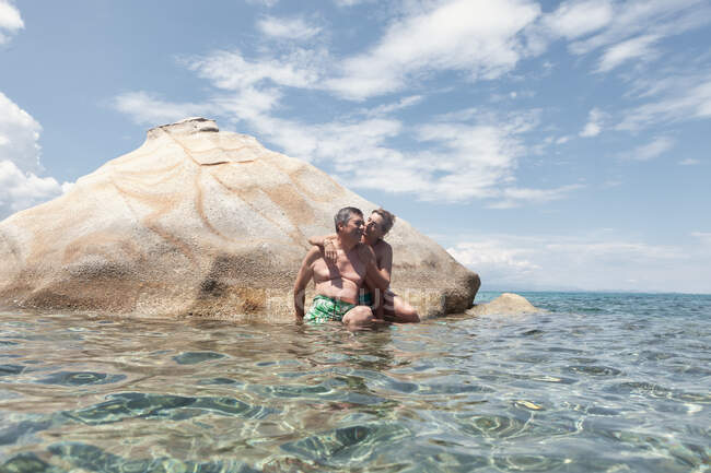 Содержание зрелый загорелый мужчина и женщина обнимая, сидя на скале в чистой воде наслаждаясь отдыхом вместе, Халкидики, Греция — стоковое фото