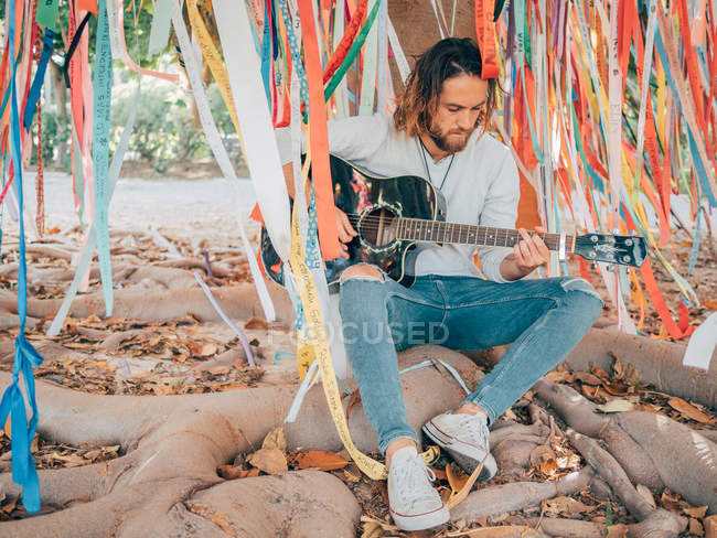 Barbudo sentado bajo el árbol decorado tocando la guitarra - foto de stock