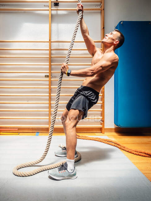 Vue latérale d'un sportif musclé torse nu montant une corde tout en faisant de l'exercice dans une salle de gym moderne — Photo de stock