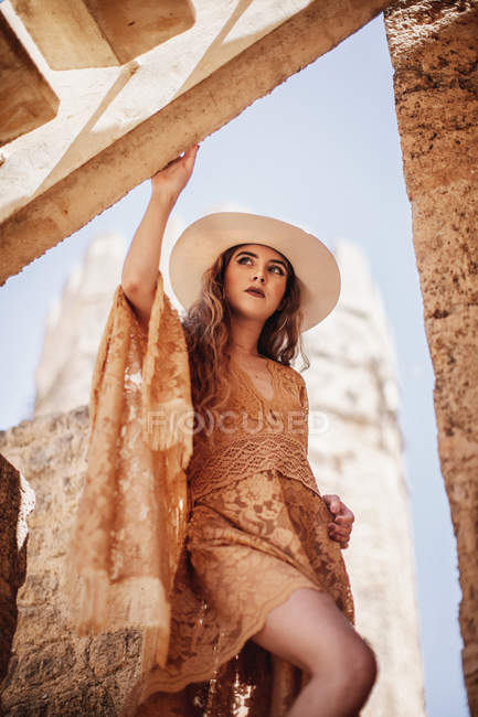 Знизу стильна жінка в елегантній бежевій сукні, що стоїть біля старої будівлі, спираючись на сходи і дивлячись вгору — стокове фото