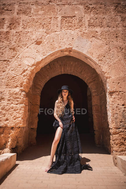 Barefoot beautiful woman in black dress and hat posing by gate of old castle on sunny day — Stock Photo