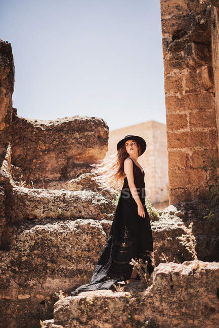 Seitenansicht einer stilvollen Frau in langem schwarzen Kleid und Hut, die neben einem zerstörten Gebäude steht und zurückblickt — Stockfoto