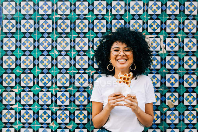 Jeune femme en t-shirt blanc debout près du mur de tuiles colorées, mangeant gaufre et souriant à la caméra — Photo de stock