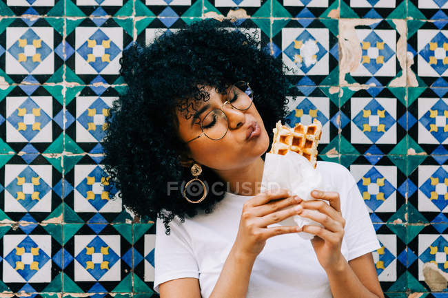 Jovem mulher em t-shirt branca em pé por parede de azulejo colorido, comendo waffle — Fotografia de Stock