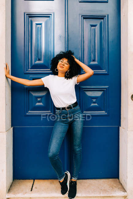 Mujer étnica bonita en camiseta blanca y jeans apoyados en la puerta azul con los ojos cerrados - foto de stock