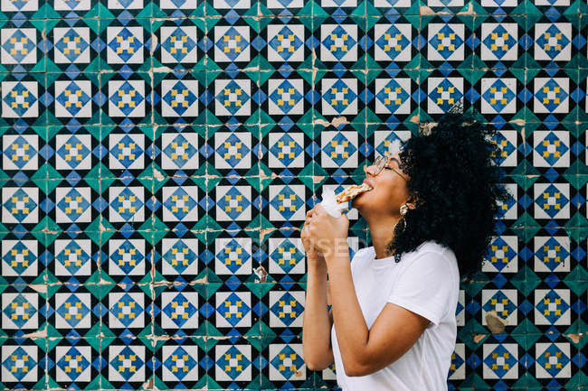 Молода жінка в білій футболці стоїть біля барвистої стіни плитки, кусає вафельку — стокове фото
