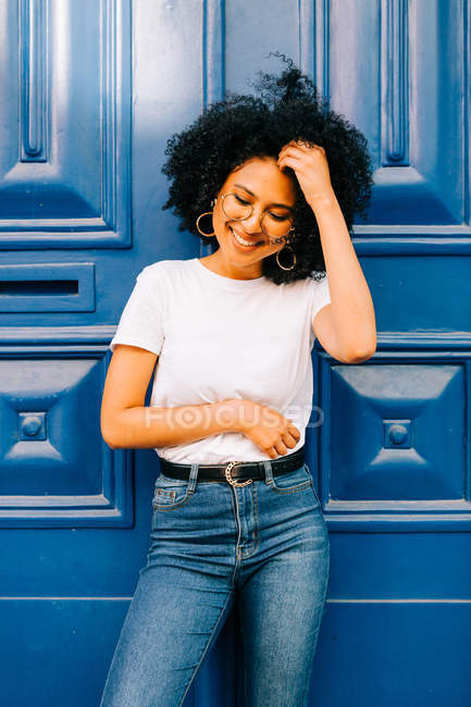 Mujer étnica bonita en camiseta blanca y jeans apoyados en la puerta azul - foto de stock