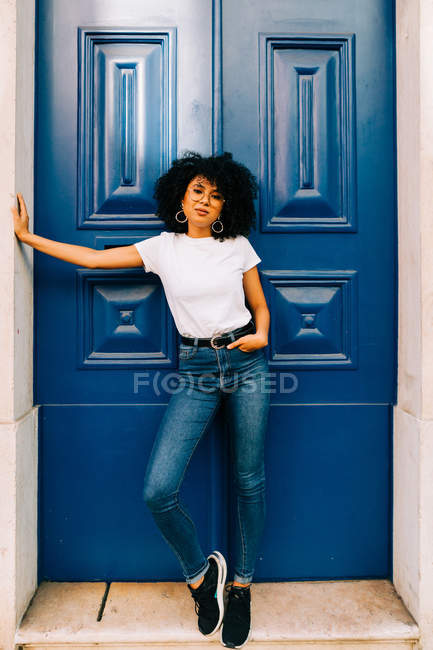 Jolie femme ethnique en t-shirt blanc et jean appuyé sur la porte bleue et regardant la caméra — Photo de stock