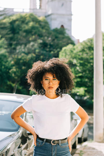Jolie femme ethnique en t-shirt blanc et aux cheveux bouclés noirs regardant la caméra sur la rue — Photo de stock
