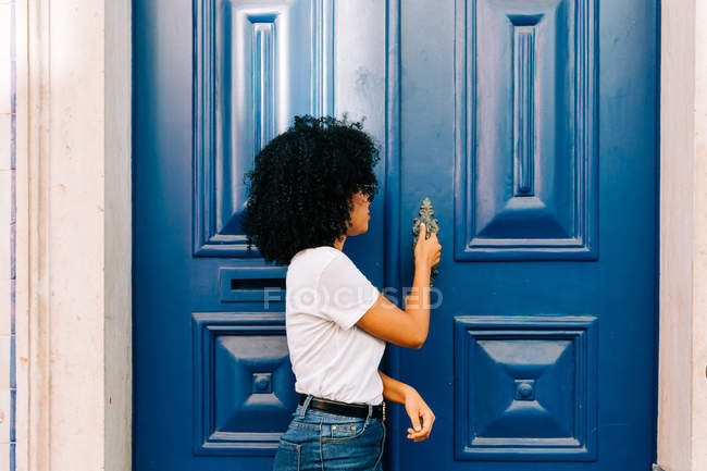 Гарненька етнічна жінка в білій футболці і джинсах стукає сині двері, дивлячись геть — стокове фото