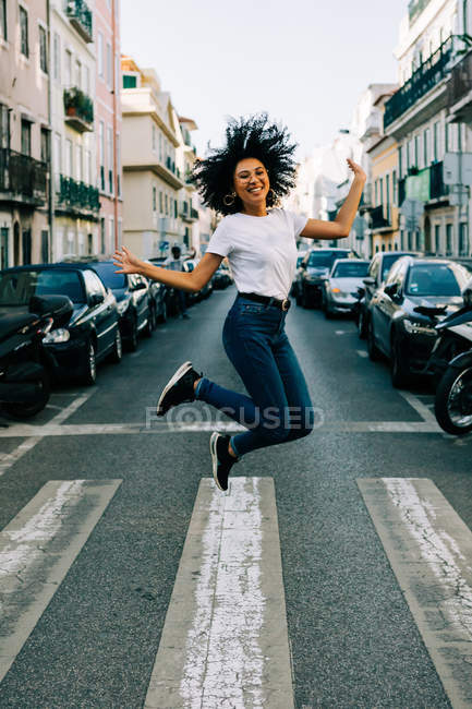 Joven mujer afroamericana alegre en jeans y camiseta blanca saltando de alegría en la calle de la ciudad durante el día - foto de stock