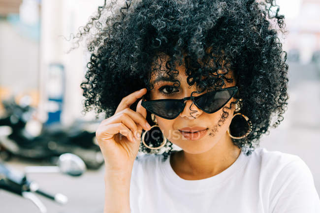 Pretty ethnic woman in white t-shirt and with black curly hair looking at  camera over black glasses — golden, gorgeous - Stock Photo | #295760004
