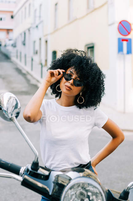 Jeune femme afro-américaine avec des cheveux bouclés noirs assis sur la moto et regardant la caméra sur les lunettes de soleil — Photo de stock