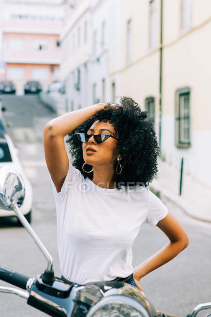 Jeune femme afro-américaine avec des cheveux bouclés noirs assis sur la moto et regardant loin sur les lunettes de soleil — Photo de stock