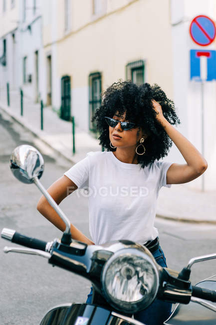 Junge afrikanisch-amerikanische Frau mit schwarzem lockigem Haar sitzt auf einem Motorrad und blickt über eine Sonnenbrille hinweg — Stockfoto