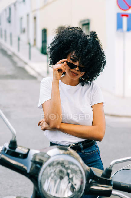 Junge afrikanisch-amerikanische Frau mit schwarzem lockigem Haar sitzt auf einem Motorrad und blickt über eine Sonnenbrille in die Kamera — Stockfoto