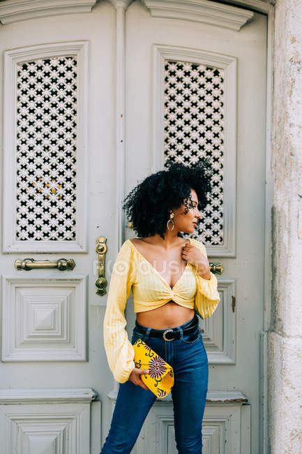 Trendy African American woman in yellow crop top and jeans standing by door and looking away — Stock Photo