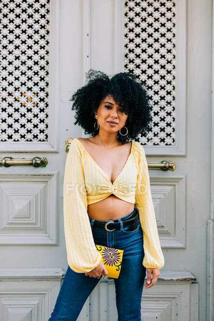 Trendige Afroamerikanerin in gelbem Crop Top und Jeans steht vor der Tür und blickt in die Kamera — Stockfoto