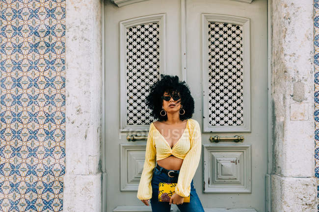 Trendy African American woman in yellow crop top and jeans standing by door and looking at camera — Stock Photo