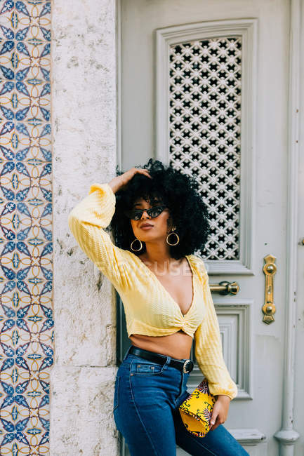 Mulher afro-americana na moda em top de cultura amarela e jeans em pé à porta e olhando para longe — Fotografia de Stock