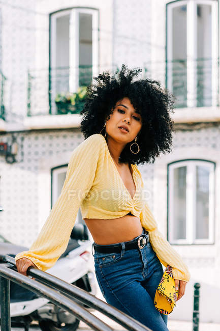 Fashionable African American woman in crop top and jeans leaning on railing and smiling — Stock Photo