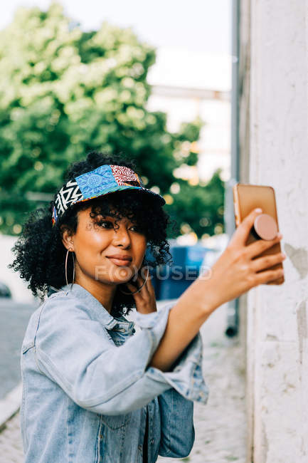 Femme afro-américaine en veste de denim et casquette colorée debout avec smartphone et prendre des photos sur la rue de la ville — Photo de stock