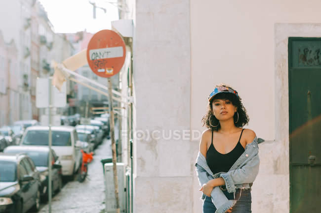 Femme afro-américaine à la mode en débardeur et veste en denim déboutonnée debout sur le trottoir et regardant la caméra — Photo de stock