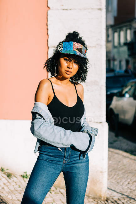 Donna alla moda in jeans e canotta in piedi e appoggiata al muro nella giornata di sole — Foto stock