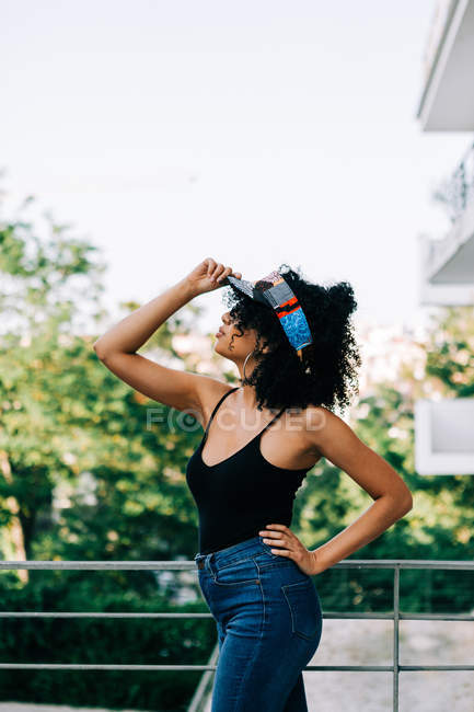 Hermosa joven afroamericana en jeans, camiseta sin mangas y diadema apoyada en barandilla y mirando hacia otro lado - foto de stock
