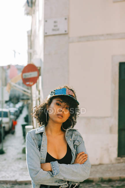 Fashionable African American woman in tank top and unbuttoned denim jacket standing on sidewalk and looking at camera — Stock Photo