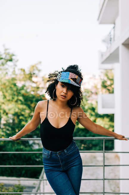 Jeune femme afro-américaine en jeans, débardeur et bandeau appuyé sur la rampe et regardant la caméra — Photo de stock