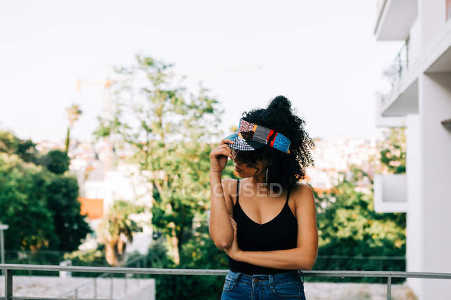 Jeune femme afro-américaine en jeans, débardeur et bandeau appuyé sur la rampe et détournant les yeux — Photo de stock