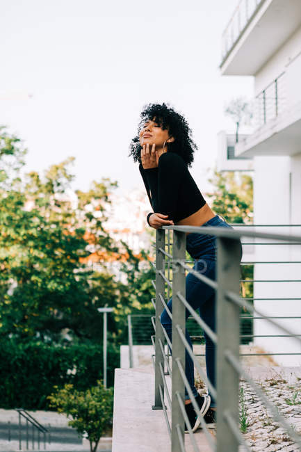 Junge Frau mit lockigem Haar steht auf Balkon, lehnt an Geländer und schaut weg — Stockfoto