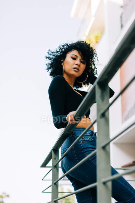 Sensual jovem mulher com cabelos cacheados em pé na varanda, inclinando-se sobre trilhos e olhando para a câmera — Fotografia de Stock