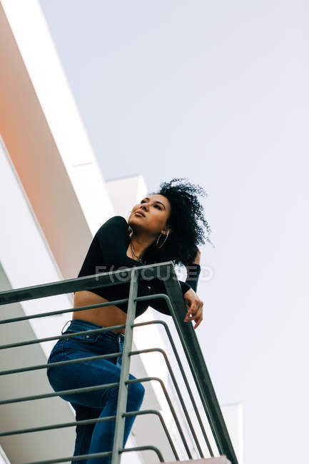 De baixo jovem mulher com cabelo encaracolado em pé na varanda, inclinando-se sobre trilhos e olhando para trás — Fotografia de Stock