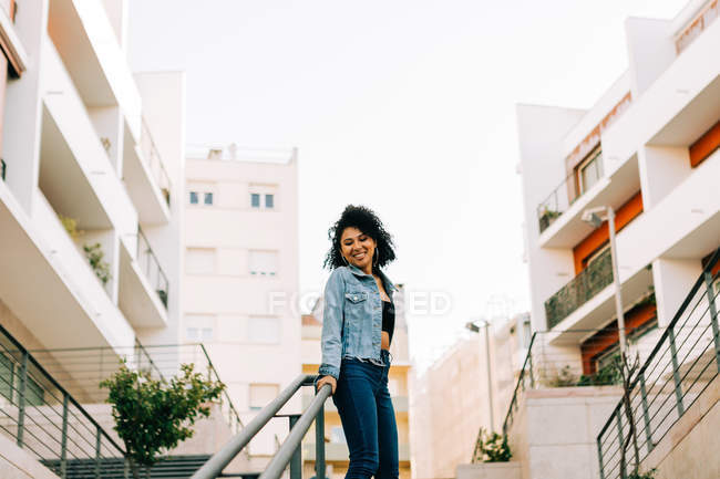 Mujer afroamericana de moda en top de cosecha y jeans apoyados en barandilla y sonriente - foto de stock