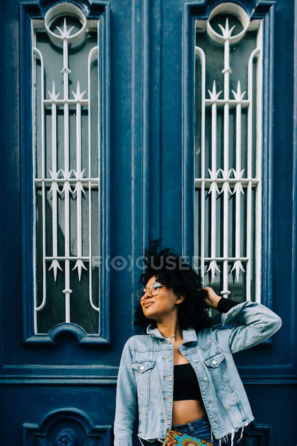 Africano mulher americana em t-shirt branca e jeans inclinado na porta azul e olhando para longe — Fotografia de Stock