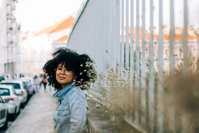 Side view of cheerful African American woman in denim jacket standing on sidewalk, leaning on fence — Stock Photo