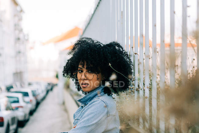 Side view of cheerful African American woman in denim jacket standing on sidewalk, leaning on fence — Stock Photo