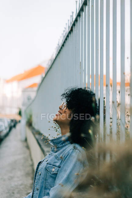 Side view of cheerful African American woman in denim jacket standing on sidewalk, leaning on fence and smiling — Stock Photo